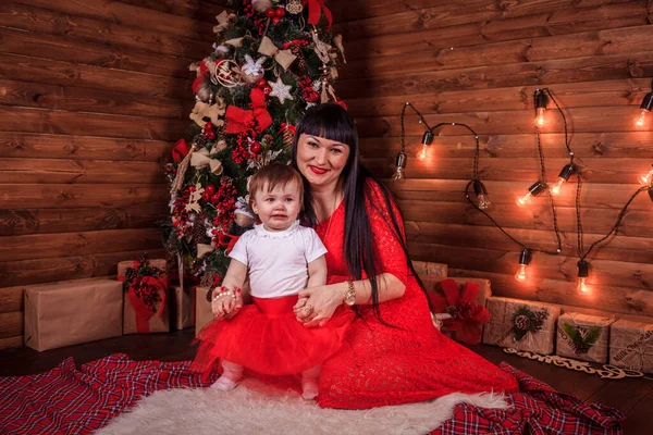 Mom and daughter are sitting under the New Year tree. Christmas family. New Year holiday. Dressed up Christmas tree. Stock Photo