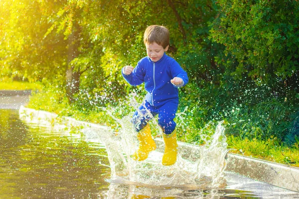 A happy boy in rubber boots jumps in puddles. The boy jumps in a puddle . Bad weather. puddles after rain. Childhood. A child in rubber boots. Summer walk. Happy child.