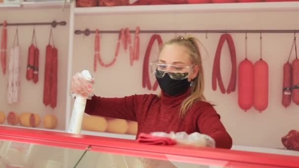 Seorang gadis bertopeng medis, kacamata dan sarung tangan khusus mendisinfeksi jendela toko. — Stok Video