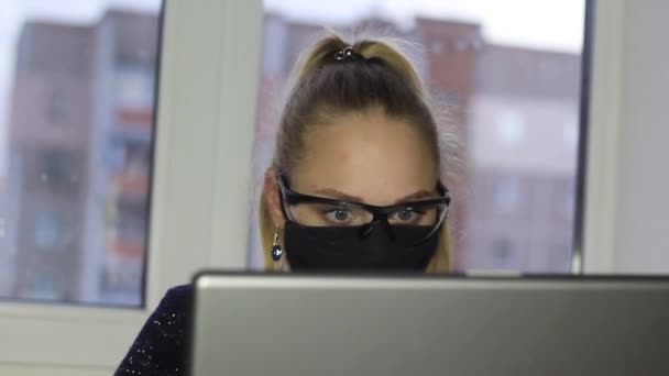 Het freelancer meisje werkt op een laptop tijdens de coronovirus epidemie in een medisch masker en bril. — Stockvideo