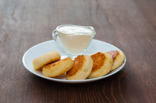 Panqueques de queso frito con crema agria en plato blanco sobre fondo de madera oscura. Para diseño de menú de cafetería o restaurante . — Foto de Stock