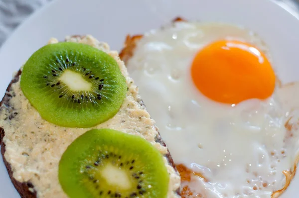 Здоровый и вкусный завтрак, сэндвич с хлебом из цельной муки с киви и жареным яйцом на белой тарелке, крупный план, вид сверху — стоковое фото