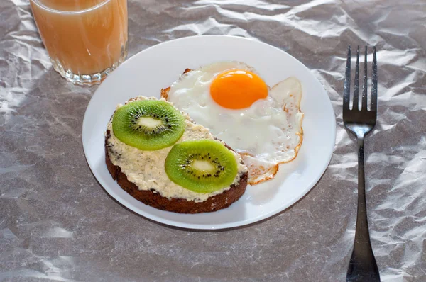 Café da manhã saudável e saboroso, suco, sanduíche integral com kiwi e ovo frito na placa branca — Fotografia de Stock