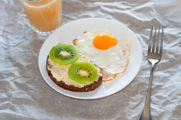 Café da manhã saudável e saboroso, suco, sanduíche integral com kiwi e ovo frito na placa branca — Fotografia de Stock