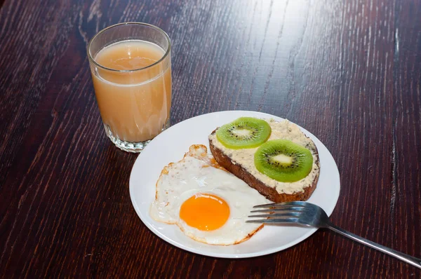 Hälsosam, välsmakande frukost, juice, fullkornsmjöl smörgås med kiwi och ett stekt ägg på en vit tallrik, på ett träbord — Stockfoto