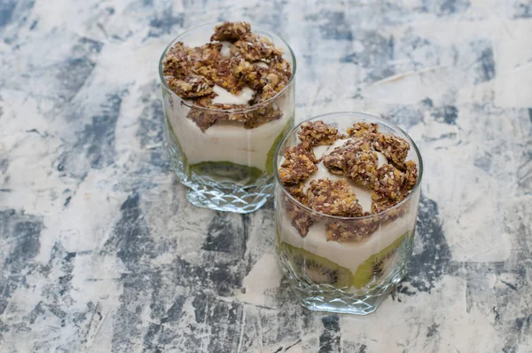 Granola, kiwi, banane et yaourt grec dans des tasses en verre sur béton gris — Photo