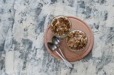 Tatlı granola, Yunan yoğurt, kivi ve muz iki cam bardak kaşık ile tahta yuvarlak tahta, gri beton, düz yatıyordu. Fitness, şekil, vücut ve sağlıklı gıda