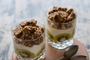 Tatlı granola, Yunan yoğurt, kivi ve muz iki cam bardak bir tahta yuvarlak tahta, gri beton, yakın çekim üzerinde kaşık ile. Fitness, şekil, vücut ve sağlıklı gıda