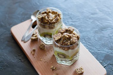 Yunan yoğurdu, granola, muz ve kivi. Ahşap platformda kaşık ile cam bardak tatlılar, koyu arka plan, üstten görünümü. Konsept vücut ve sağlıklı gıda