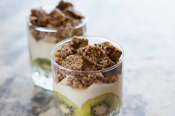 Granola, kiwi, banana and Greek yogurt in two glass cups on gray concrete, copy space. Fitness, figure, body and healthy food — Stock Photo, Image