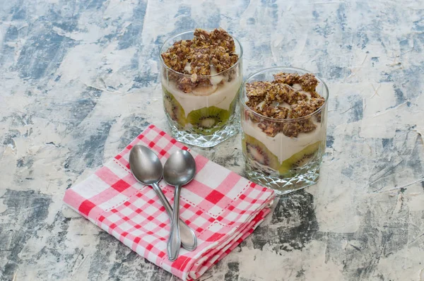 Dessert granola, Greek yogurt, kiwi and banana in two glass cups with spoons and pink checkered towel on gray concrete. Fitness, figure, body and healthy food