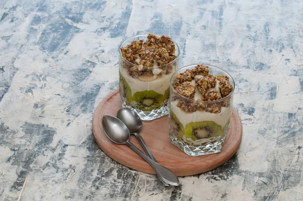 Dessert granola, Greek yogurt, kiwi and banana in two glass cups with spoons on wooden round board, gray concrete. Fitness, figure, body and healthy food