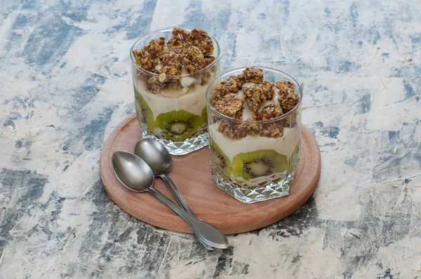 Dessert granola, Greek yogurt, kiwi and banana in two glass cups with spoons on wooden round board, gray concrete. Fitness, figure, body and healthy food