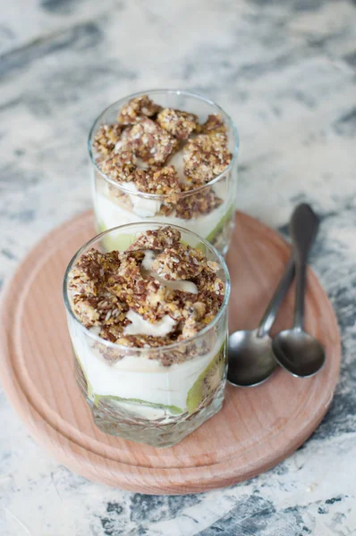 Dessert granola, greek yogurt, kiwi and banana in two glass cups with spoons on a wooden round board, gray concrete, close-up vertical frame format. Fitness, figure, body and healthy food