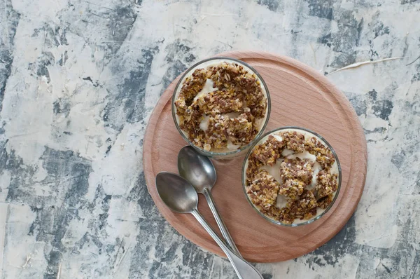 Dessert granola, Greek yogurt, kiwi and banana in two glass cups with spoons on a wooden round board, gray concrete, flat lay. Fitness, figure, body and healthy food