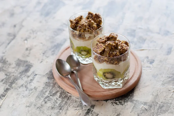 Dessert granola, Greek yogurt, kiwi and banana in two glass cups with spoons on wooden round board, gray concrete, close-up. Fitness, figure, body and healthy food