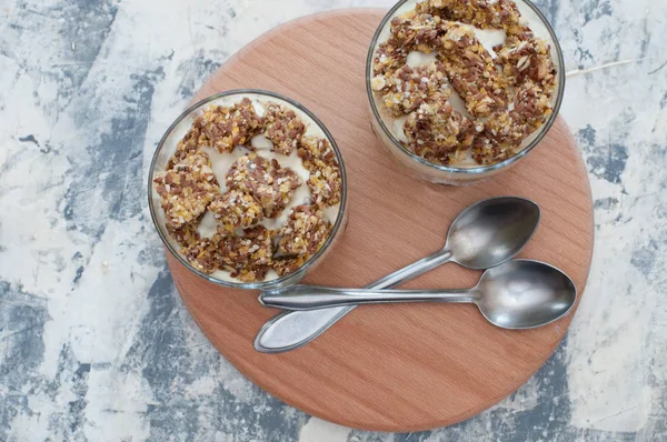 Dessert granola, Greek yogurt, kiwi and banana in two glass cups with spoons on a wooden round board, gray concrete, copy space, flat lay. Fitness, figure, body and healthy food