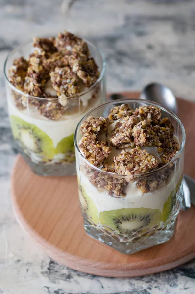 Dessert granola, Greek yogurt, kiwi and banana in two glass cups with spoons on a wooden round board, gray concrete, copy space, vertical format. Fitness, figure, body and healthy food