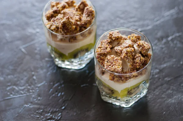 Yaourt grec, granola, banane et kiwi. Deux desserts dans des tasses en verre sur fond sombre. Concept corps et alimentation saine — Photo
