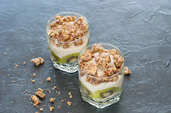 Yaourt grec, granola, banane et kiwi. Desserts dans des tasses en verre sur fond sombre, vue latérale du haut. Concept corps et alimentation saine — Photo