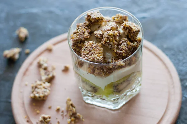 Granola, banane, kiwi et yaourt grec. Dessert dans une tasse en verre sur une planche ronde en bois, fond sombre. Concept corps et alimentation saine — Photo