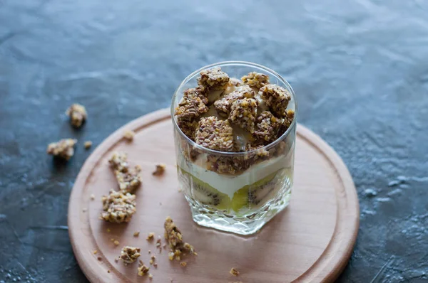 Granola, banan, kiwi och grekisk yoghurt. Dessert i Glaskopp på en trä rund bräda, mörk bakgrund. Koncept kropp och hälsosam mat — Stockfoto