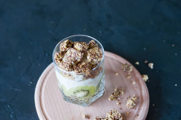 Granola, banana, kiwi and greek yogurt. Dessert in glass cup on a wooden round board, dark background. Concept body and healthy food — Stock Photo, Image