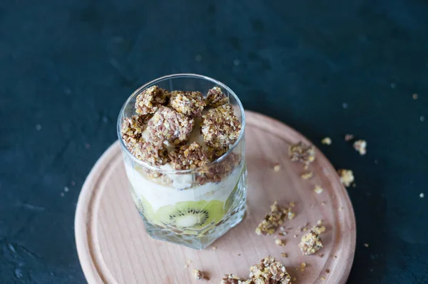 Granola, banana, kiwi e iogurte grego. Sobremesa em copo de vidro em uma tábua redonda de madeira, fundo escuro. Conceito corpo e alimentos saudáveis — Fotografia de Stock