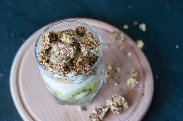 Granola, banane, kiwi et yaourt grec. Dessert dans une tasse en verre sur une planche ronde en bois, fond sombre. Concept corps et alimentation saine — Photo