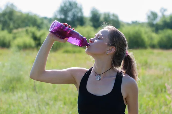 Joven hermosa mujer muy bonita en un traje deportivo bebe agua de una botella después del entrenamiento en yoga, gimnasia al aire libre en el verano —  Fotos de Stock