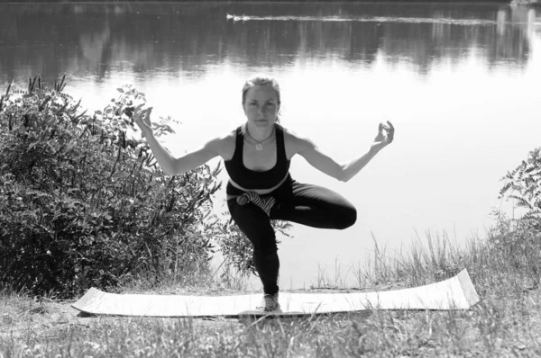 Menina bonito executa asanas ioga na margem de um rio, lago ou mar em uma manhã de verão ensolarada. Dieta, exercício e ioga na natureza, bw — Fotografia de Stock