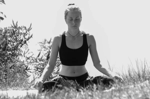 Menina bonito executa asanas ioga na margem de um rio, lago ou mar em uma manhã de verão ensolarada. Dieta, exercício e ioga na natureza, bw — Fotografia de Stock