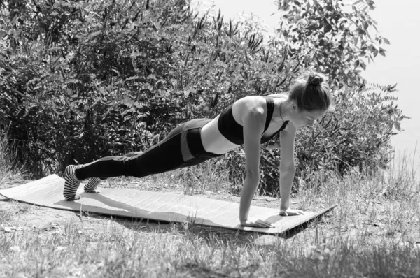 Menina bonito executa asanas ioga na margem de um rio, lago ou mar em uma manhã de verão ensolarada. Dieta, exercício e ioga na natureza, bw — Fotografia de Stock