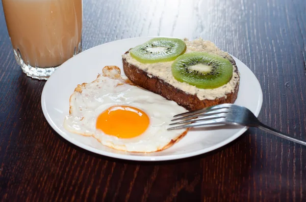 Здоровый, вкусный завтрак, сок, сэндвич с хлебом из цельной муки с киви и яичницей на белой тарелке, на деревянном столе — стоковое фото