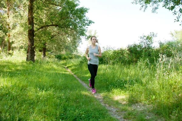 Mujer hermosa, atlética, delgada con auriculares y traje deportivo corre por el parque, verano, mañana soleada. Correr por la mañana y deportes — Foto de Stock