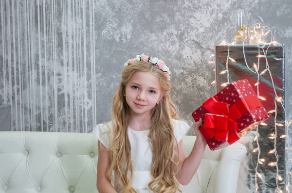 Une belle fille aux cheveux blonds et aux cheveux longs dans une robe blanche tient une boîte avec un cadeau. L'enfant est assis sur un canapé blanc près de la guirlande de Noël — Photo