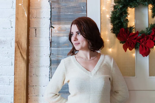 A young woman stands at the door of the house decorated with a garland and a New Year\'s wreath of fir pine branches, looking away, dreaming