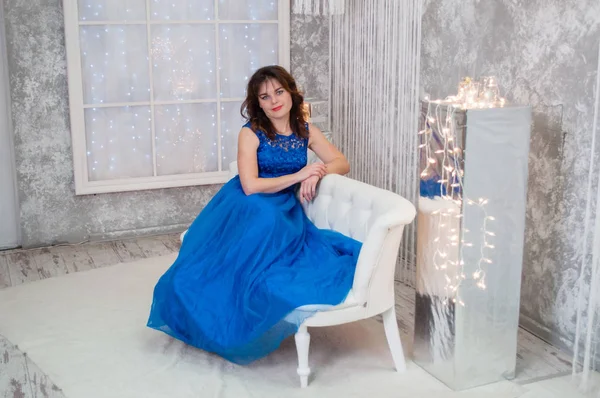 Mulher de cabelos castanhos muito bonita com cabelo encaracolado em um vestido de noite azul sentado em um sofá perto da grinalda do Ano Novo — Fotografia de Stock