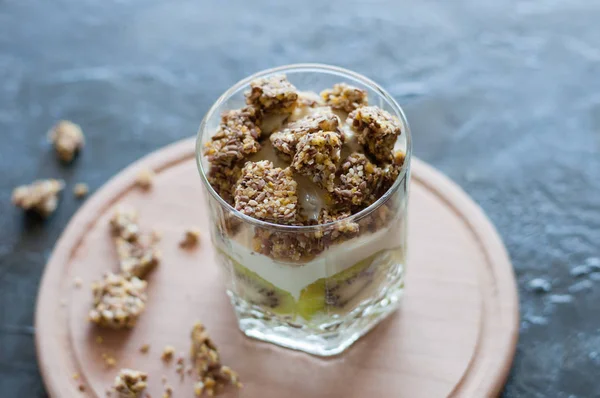 Granola, banane, kiwi et yaourt grec. Dessert dans une tasse en verre sur une planche ronde en bois, fond sombre. Concept corps et alimentation saine — Photo