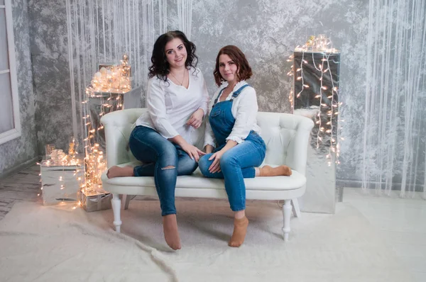 Duas meninas bonitas celebram o Natal, Ano Novo. Meninas no interior do Ano Novo da sala estão sentados em um sofá branco contra o fundo de guirlandas — Fotografia de Stock