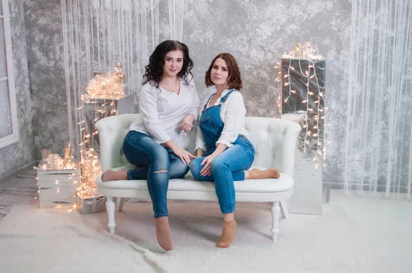 Duas meninas bonitas celebram o Natal, Ano Novo. Meninas no interior do Ano Novo da sala estão sentados em um sofá branco contra o fundo de guirlandas — Fotografia de Stock