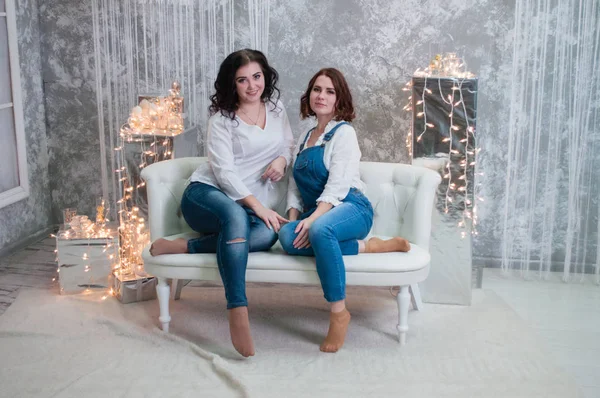 Duas meninas bonitas celebram o Natal, Ano Novo. Meninas no interior do Ano Novo da sala estão sentados em um sofá branco contra o fundo de guirlandas — Fotografia de Stock