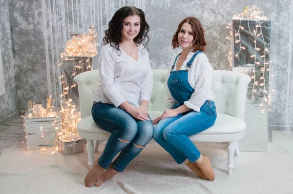 Duas meninas muito bonitas celebram o Natal, ano novo. Meninas no interior do Ano Novo da sala estão sentados em um sofá branco contra o fundo de guirlandas — Fotografia de Stock