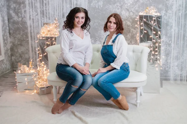 Duas meninas bonitas celebram o Natal, Ano Novo. Meninas no interior do Ano Novo da sala estão sentados em um sofá branco contra o fundo de guirlandas — Fotografia de Stock