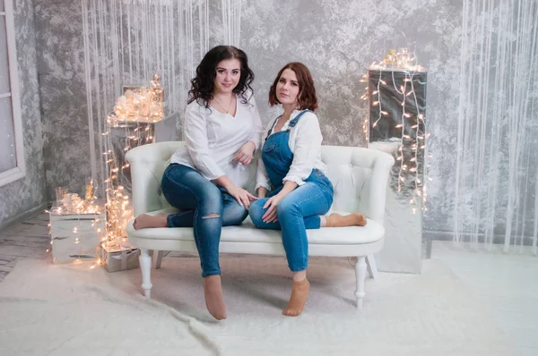 Due belle ragazze festeggiano il Natale, Capodanno. Le ragazze all'interno della stanza del nuovo anno sono sedute su un divano bianco sullo sfondo delle ghirlande — Foto Stock