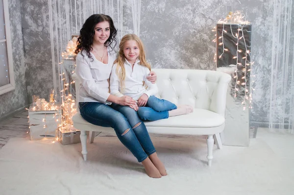 Due ragazze molto carine, le sorelle festeggiano il Natale, il nuovo anno. Le ragazze all'interno della stanza del nuovo anno sono sedute su un divano bianco sullo sfondo delle ghirlande — Foto Stock