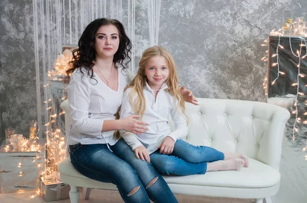Due ragazze molto carine, le sorelle festeggiano il Natale, il nuovo anno. Le ragazze all'interno della stanza del nuovo anno sono sedute su un divano bianco sullo sfondo delle ghirlande — Foto Stock