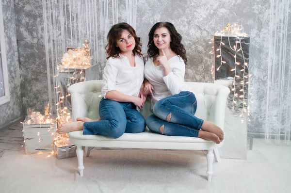 Duas jovens muito bonitas celebram o Natal, o ano novo. Meninas no interior do Ano Novo da sala estão sentados em um sofá branco contra o fundo de guirlandas — Fotografia de Stock