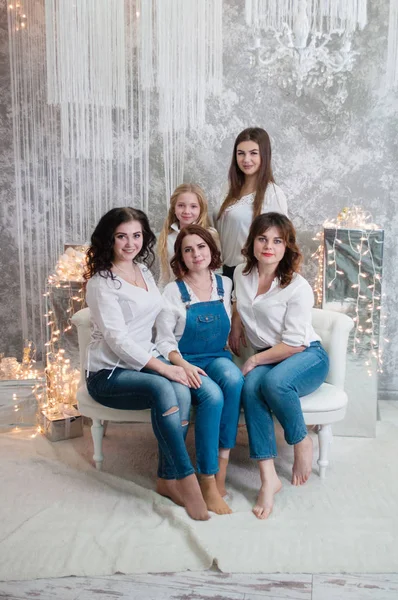 La compagnia di belle ragazze festeggia il Natale, il nuovo anno. Le ragazze all'interno della stanza del nuovo anno sono sedute su un divano bianco sullo sfondo delle ghirlande — Foto Stock