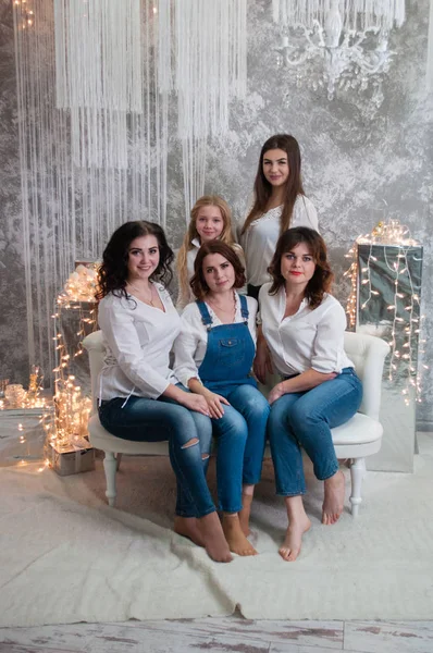 La compagnia di belle ragazze festeggia il Natale, il nuovo anno. Le ragazze all'interno della stanza del nuovo anno sono sedute su un divano bianco sullo sfondo delle ghirlande — Foto Stock